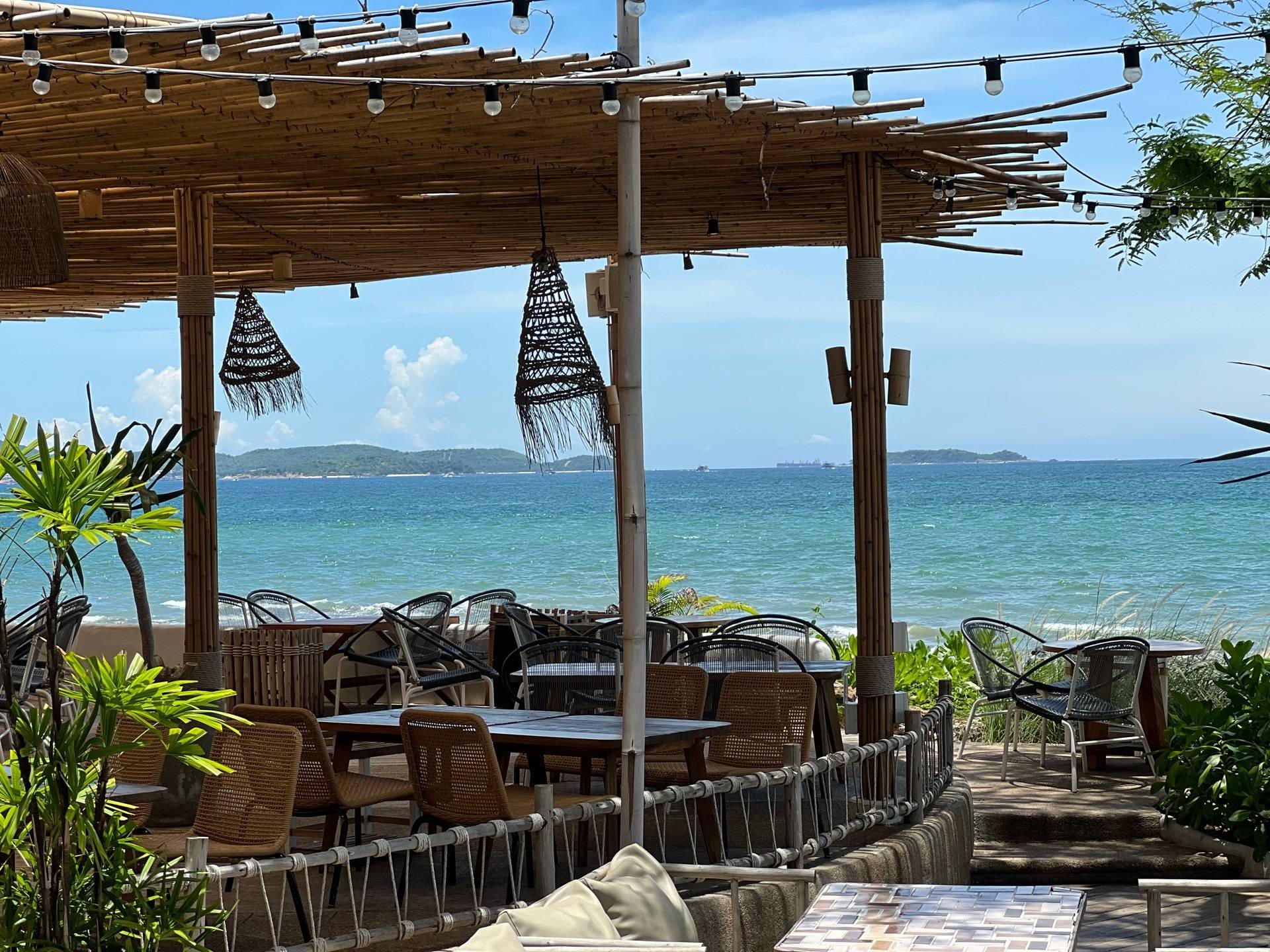  A bamboo-covered patio with tables and chairs sits directly on a beach, overlooking the ocean on a sunny day.