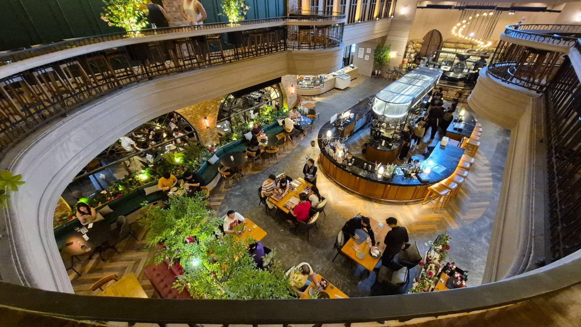 A view from a balcony overlooking a large, two-story restaurant with a busy central bar and people dining at tables.